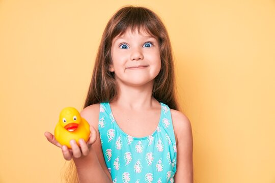 Little caucasian kid girl with long hair wearing swimsuit and holding duck toy looking positive and happy standing and smiling with a confident smile showing teeth