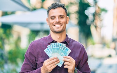 Young hispanic man smiling happy holding brazilian real banknotes at the city.