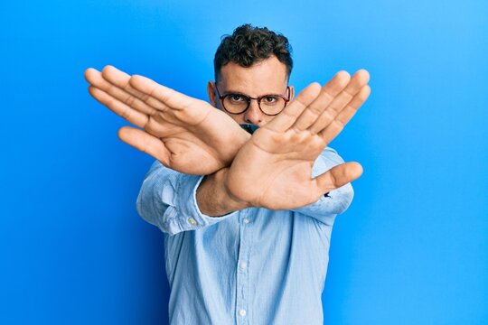 Young hispanic man wearing casual clothes and glasses rejection expression crossing arms and palms doing negative sign, angry face