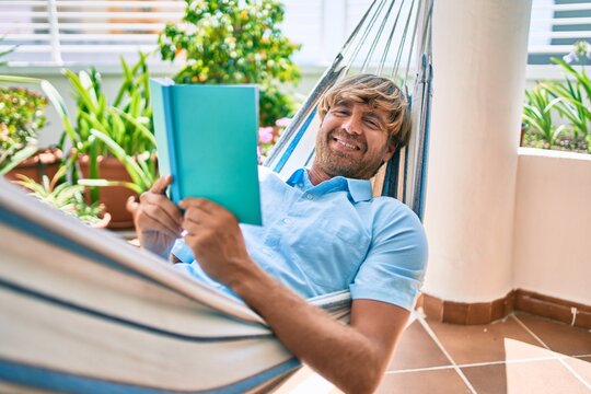 Middle age handsome man at the terrace of his house relaxing lying on a hammock reading a book