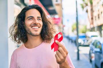 Young hispanic man smiling happy holding hiv awareness red ribbon at city.