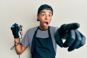 Young handsome african american man tattoo artist wearing professional uniform and gloves holding tattooer machine pointing with finger surprised ahead, open mouth amazed expression.