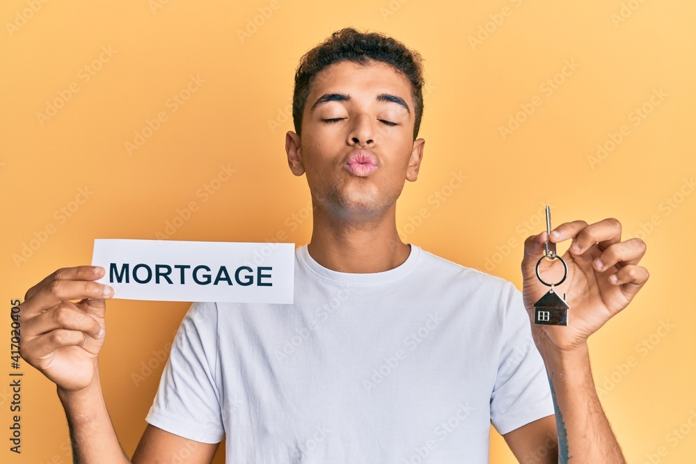 Canvas Prints Young handsome african american man holding paper with mortgage word and house keys looking at the camera blowing a kiss being lovely and sexy. love expression.