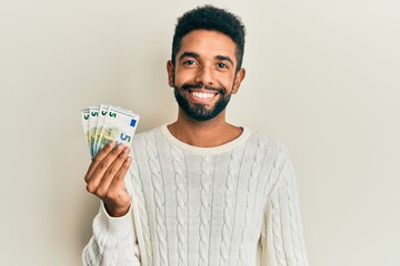 Handsome hispanic man with beard holding bunch of 5 euro banknotes looking positive and happy...