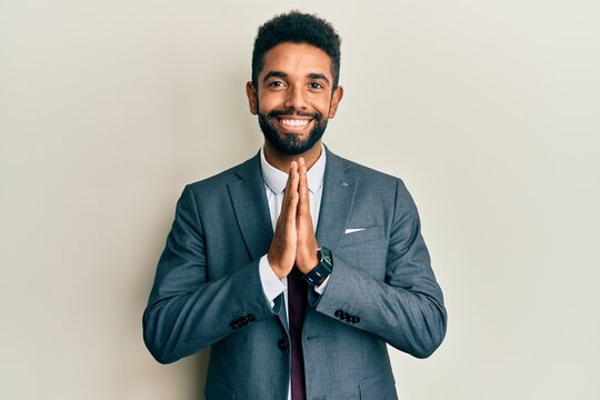 Handsome hispanic man with beard wearing business suit and tie praying with hands together asking for forgiveness smiling confident.