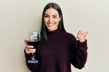 Young brunette woman drinking a glass of red wine pointing thumb up to the side smiling happy with open mouth