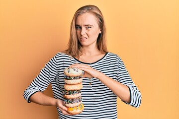 Beautiful blonde woman holding tasty colorful doughnuts clueless and confused expression. doubt...