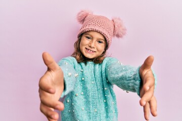 Little beautiful girl wearing wool sweater and cute winter hat looking at the camera smiling with open arms for hug. cheerful expression embracing happiness. - obrazy, fototapety, plakaty