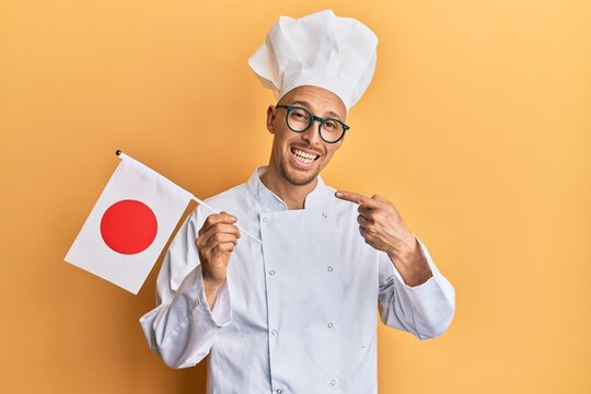 Bald man with beard wearing professional cook uniform holding japan flag smiling happy pointing with hand and finger