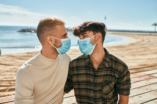 Young gay couple wearing medical mask sitting on the bench at the beach.