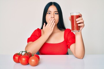 Young beautiful asian girl drinking glass of healthy tomato juice covering mouth with hand, shocked...