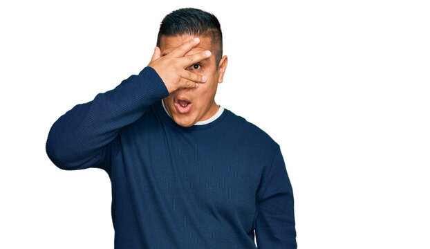Young latin man wearing casual clothes peeking in shock covering face and eyes with hand, looking through fingers with embarrassed expression.