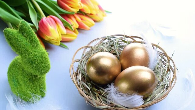 Egg color. Happy Easter decoration: golden colour eggs in basket with spring tulips, white feathers on pastel blue background. Foil minimalist egg design, modern template