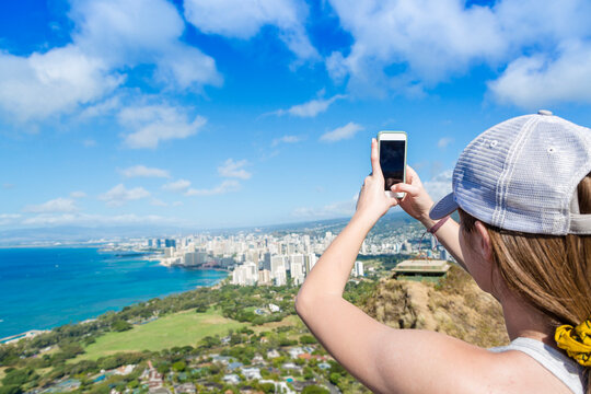 Woman Taking a cell phone photo of the city of Honolulu, Hawaii from high above the city at Diamond Head crater. Tourist enjoying the sights on her Hawaiian vacation