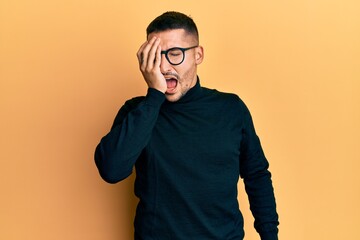 Handsome man with tattoos wearing turtleneck sweater and glasses yawning tired covering half face, eye and mouth with hand. face hurts in pain.