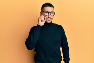 Handsome man with tattoos wearing turtleneck sweater and glasses mouth and lips shut as zip with...