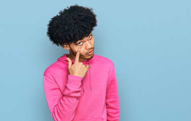 Obraz na płótnie Canvas Young african american man with afro hair wearing casual pink sweatshirt pointing to the eye watching you gesture, suspicious expression