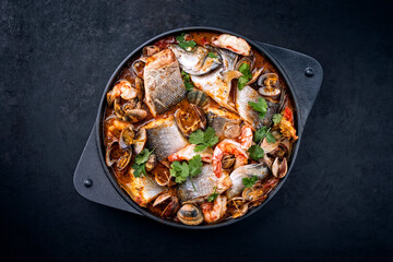 Modern style traditional Spanish seafood zarzuela de pescado with fish, king prawns and venus clams served in clear sauce as top view in design pot with copy space