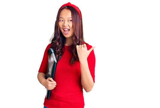 Young beautiful chinese girl wearing student folder pointing thumb up to the side smiling happy with open mouth