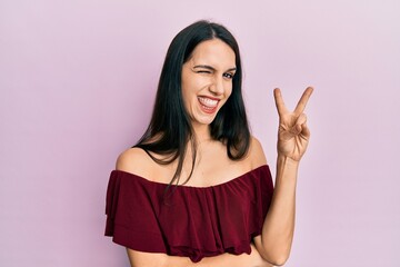 Young hispanic woman wearing casual clothes smiling with happy face winking at the camera doing victory sign. number two.