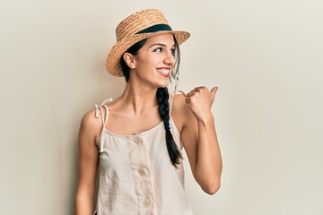 Young hispanic woman wearing summer hat pointing thumb up to the side smiling happy with open mouth