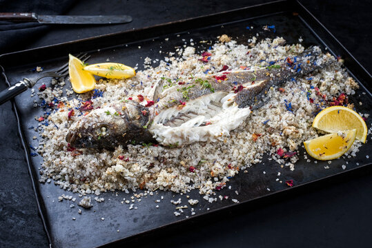 Traditional Atlantic Loup de Mer bedded into sea salt served with lemon slices and spices as close-up on a rustic metal tray