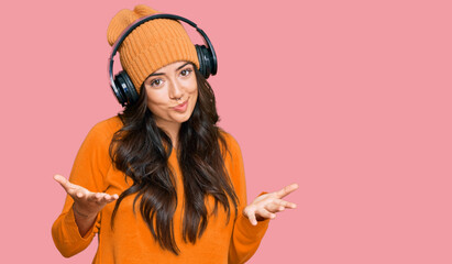 Beautiful brunette young woman listening to music using headphones clueless and confused expression with arms and hands raised. doubt concept.