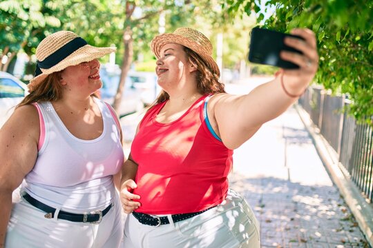 Two plus size overweight sisters twins women smiling taking a selfie picture with the phone outdoors on a sunny day