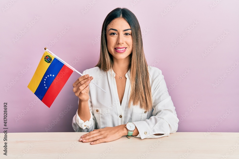 Wall mural young hispanic woman holding venezuelan flag sitting on the table looking positive and happy standin