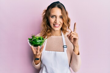 Young caucasian woman wearing apron holding green peppers bowl smiling with an idea or question pointing finger with happy face, number one