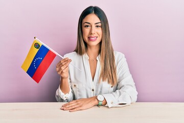 Young hispanic woman holding venezuelan flag sitting on the table looking positive and happy standing and smiling with a confident smile showing teeth