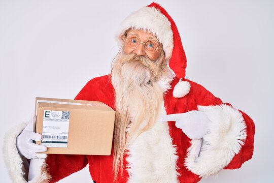 Old senior man with grey hair and long beard wearing santa claus costume holding delivery box smiling happy pointing with hand and finger