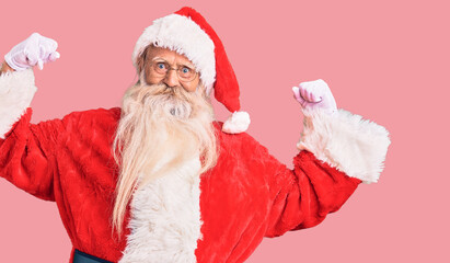 Old senior man with grey hair and long beard wearing traditional santa claus costume showing arms muscles smiling proud. fitness concept.