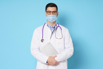 Young male doctor wearing medical mask, glasses and stethoscope round his neck, holding tablet, isolated on blue background