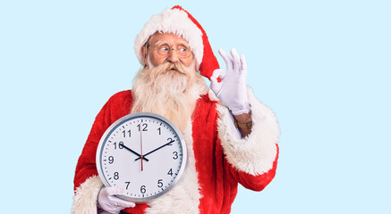 Old senior man with grey hair and long beard wearing santa claus costume holding clock doing ok sign with fingers, smiling friendly gesturing excellent symbol