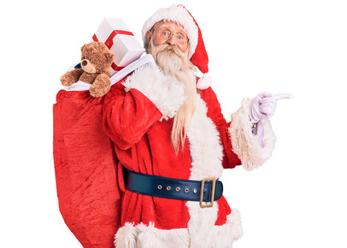 Old senior man with grey hair and long beard wearing santa claus costume holding bag with presents smiling happy pointing with hand and finger to the side