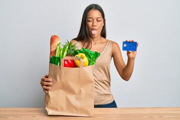 Beautiful hispanic woman holding groceries and credit card sticking tongue out happy with funny expression.