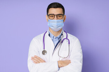 Fototapeta premium Young male doctor wearing medical mask, standing with crossed arms in white coat, isolated on purple background