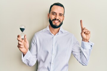 Young man with beard holding sand clock smiling with an idea or question pointing finger with happy face, number one