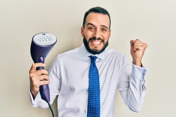 Naklejka premium Young business man with beard holding electric steam iron screaming proud, celebrating victory and success very excited with raised arm