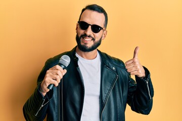 Young man with beard singing song using microphone smiling happy and positive, thumb up doing excellent and approval sign
