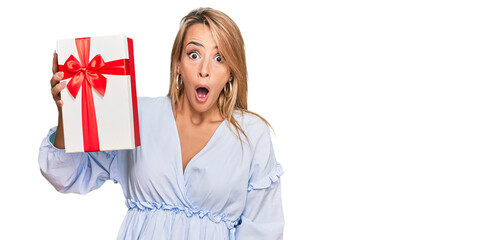 Beautiful blonde woman holding gift scared and amazed with open mouth for surprise, disbelief face