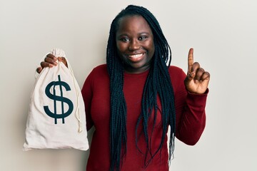 Young black woman with braids holding dollars bag smiling with an idea or question pointing finger...