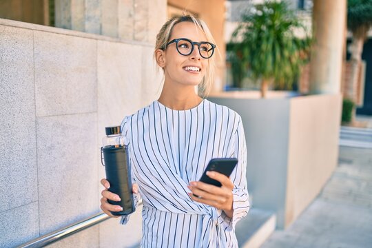 Young blonde businesswoman smiling happy using smartphone and drinking bottle of water at the city.