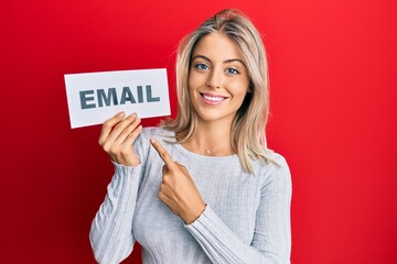 Beautiful blonde woman holding paper with email address smiling happy pointing with hand and finger