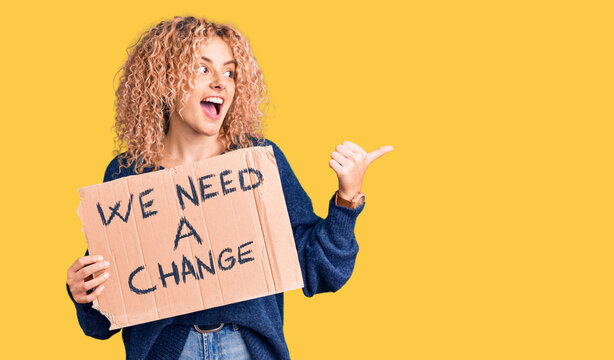 Young blonde woman with curly hair holding we need a change banner pointing thumb up to the side smiling happy with open mouth