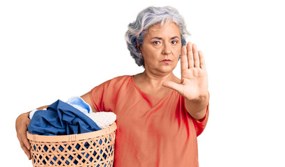 Senior woman with gray hair holding laundry basket with open hand doing stop sign with serious and confident expression, defense gesture