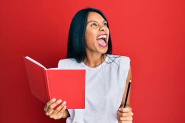 Young african american woman holding book and pencil angry and mad screaming frustrated and furious, shouting with anger. rage and aggressive concept.