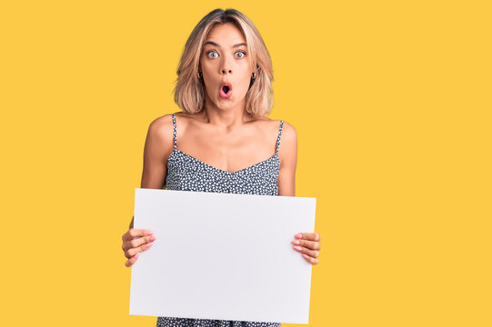 Beautiful caucasian woman holding blank empty banner scared and amazed with open mouth for surprise, disbelief face