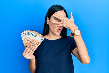 Beautiful young woman holding bunch of 50 euro banknotes peeking in shock covering face and eyes with hand, looking through fingers with embarrassed expression.